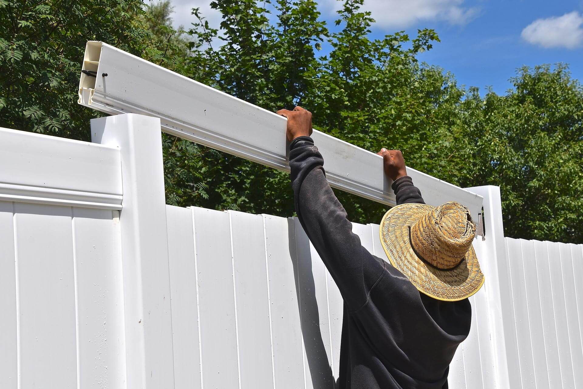 Man builds a white vinyl fence while wearing a sun hat